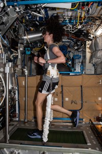 NASA Astronaut Jasmin Moghbeli runs on a treadmill while wearing a bungee harness that prevents her from floating away. Moghbeli seems focused while her hair floats above her. Several cords are near the ceiling.