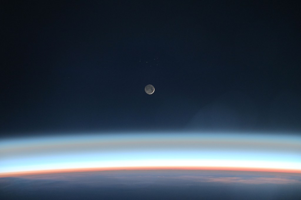 the Moon is in a waxing crescent phase, with only a small sliver of the Sun-illuminated side visible on its right side. The rest of the Moon glows, albeit less brightly, by the light of the Sun after it has been reflected off of the Earth. Below the Moon, Earth’s bright horizon arcs across the bottom of the image. It shows a cross-section of the planet’s atmospheric layers including the orange troposphere and the light-blue stratosphere.