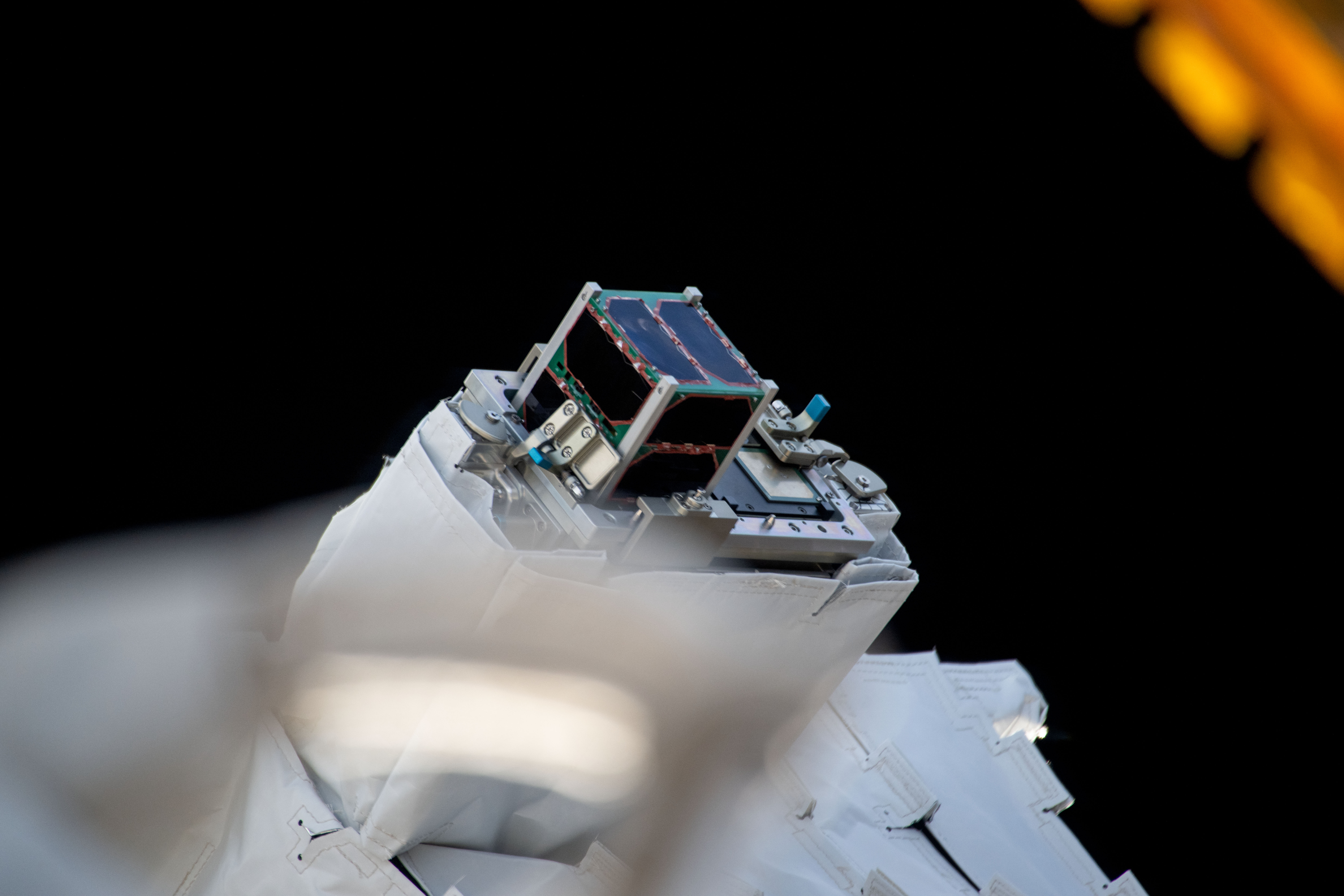 In the center, a CubeSat is attached to the outside of station preparing for deployment. Parts of space station can be seen in the bottom third of the image, while the top third shows the blackness of space.