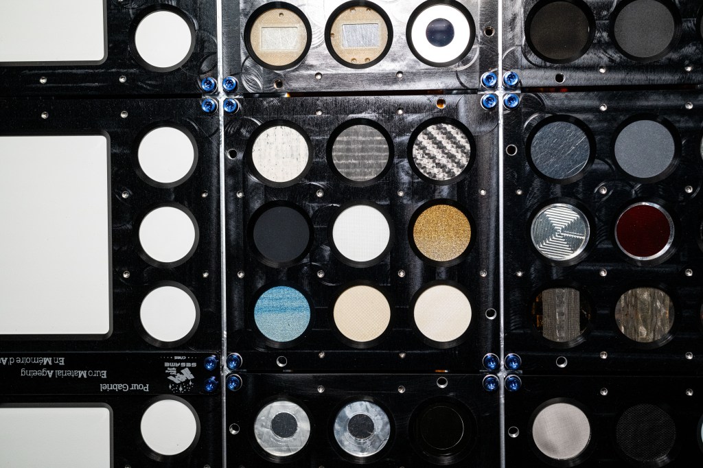 Close-up view of an experimental setup featuring an array of circular material samples mounted in a black panel with metal screws. The samples include various reflective, metallic, and translucent materials, such as silver, gold, and transparent surfaces. This experiment is designed to test material durability and properties in the harsh space conditions.