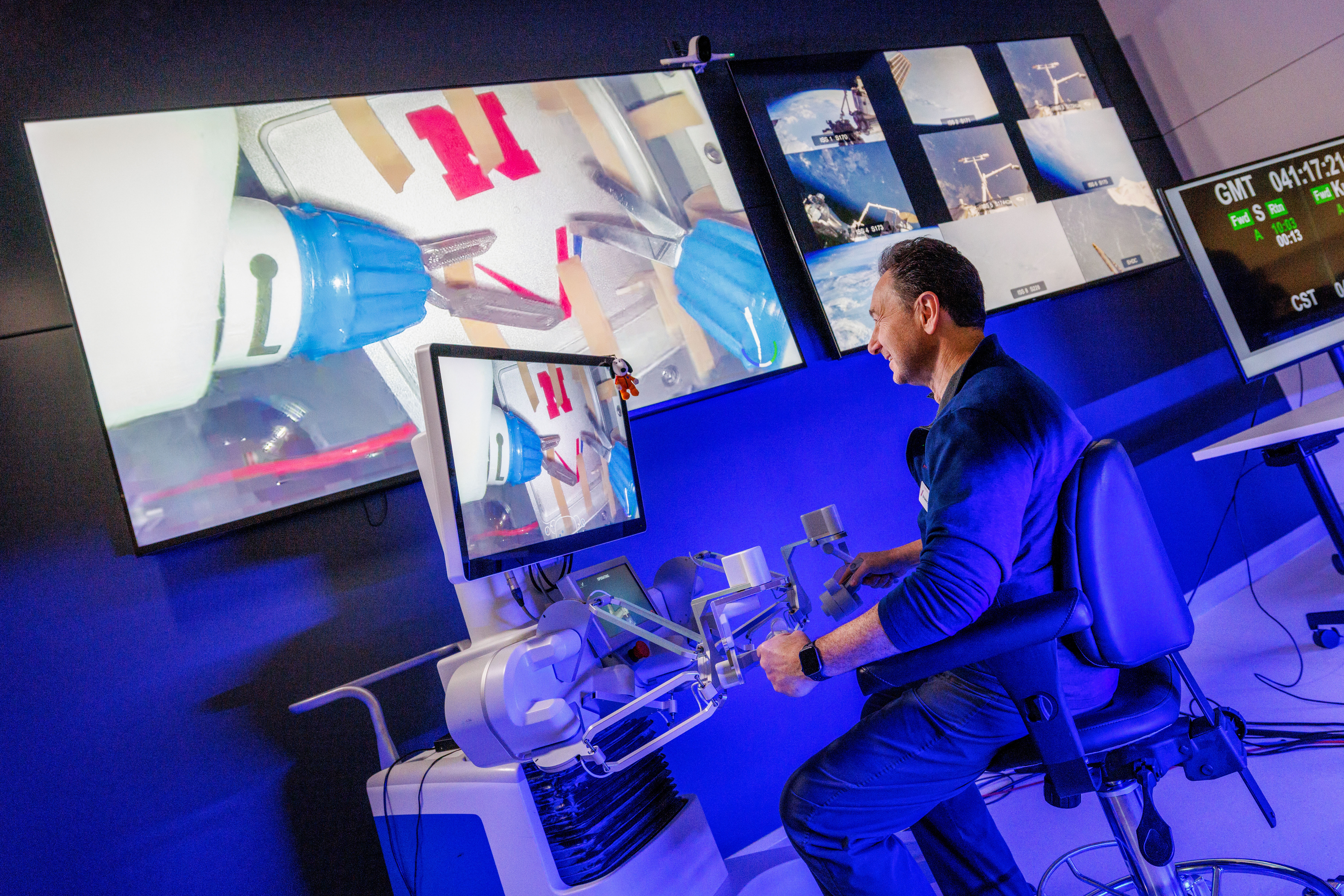 Oleynikov, wearing a blue shirt and pants, sits in a padded chair facing a computer screen on a large stand and holding in each hand controllers at the end of maneuvering arms attached to the stand. The image on the screen is projected on a larger screen on the wall.