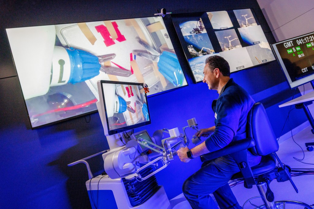 Oleynikov, wearing a blue shirt and pants, sits in a padded chair facing a computer screen on a large stand and holding in each hand controllers at the end of maneuvering arms attached to the stand. The image on the screen is projected on a larger screen on the wall.