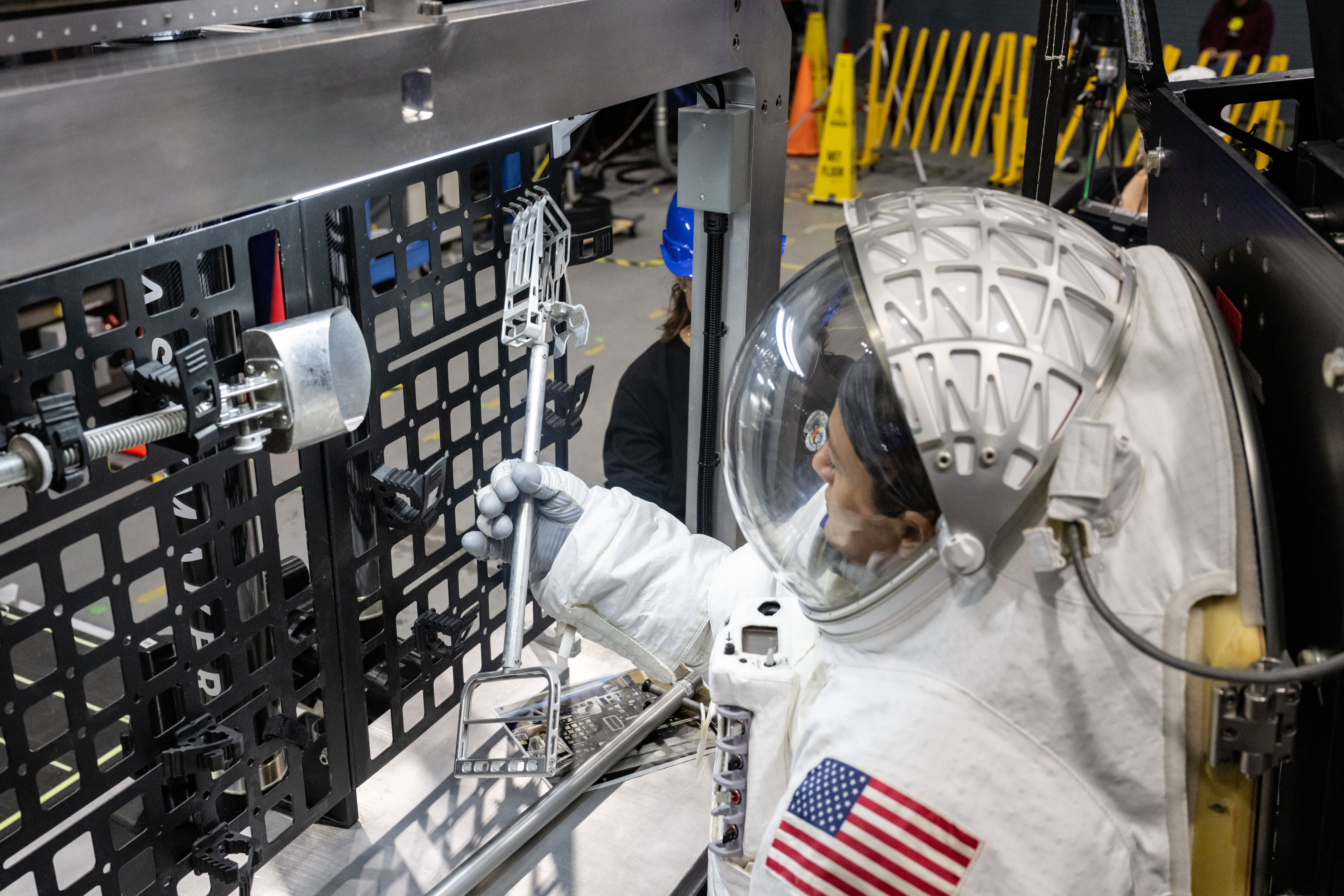 NASA astronaut Jessica Meir is shown in a white spacesuit with the American flag on her left shoulder as she grabs a lunar geology tool from a tool...