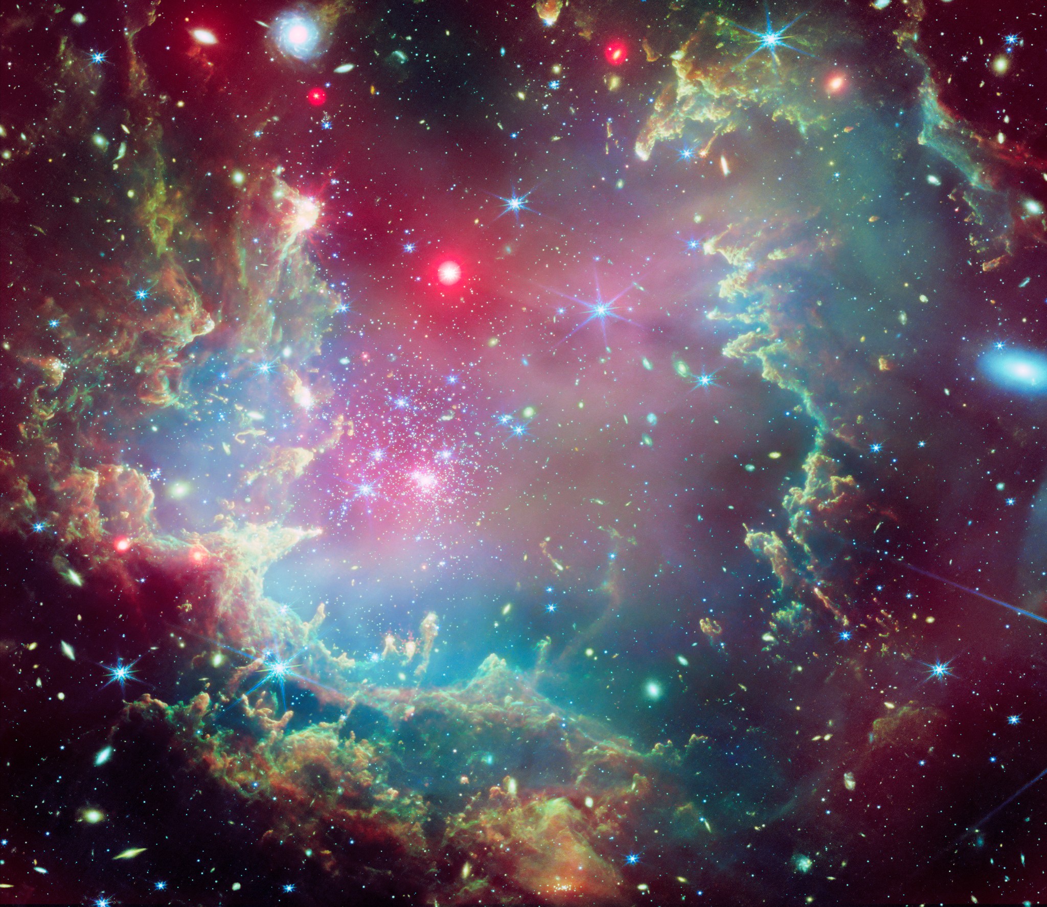Near the outskirts of the Small Magellanic Cloud, a satellite galaxy roughly 200 000 light-years from Earth, lies the young star cluster NGC 602, which is featured in this new image from the NASA/ESA/CSA James Webb Space Telescope. This image includes data from Webb’s NIRCam (Near-InfraRed Camera) and MIRI (Mid-InfraRed Instrument). The local environment of this cluster is a close analogue of what existed in the early Universe, with very low abundances of elements heavier than hydrogen and helium. The existence of dark clouds of dense dust and the fact that the cluster is rich in ionised gas also suggest the presence of ongoing star formation processes. This cluster provides a valuable opportunity to examine star formation scenarios under dramatically different conditions from those in the solar neighbourhood. [Image description: A star cluster is shown inside a large nebula of many-coloured gas and dust. The material forms dark ridges and peaks of gas and dust surrounding the cluster, lit on the inner side, while layers of diffuse, translucent clouds blanket over them. Around and within the gas, a huge number of distant galaxies can be seen, some quite large, as well as a few stars nearer to us which are very large and bright.]