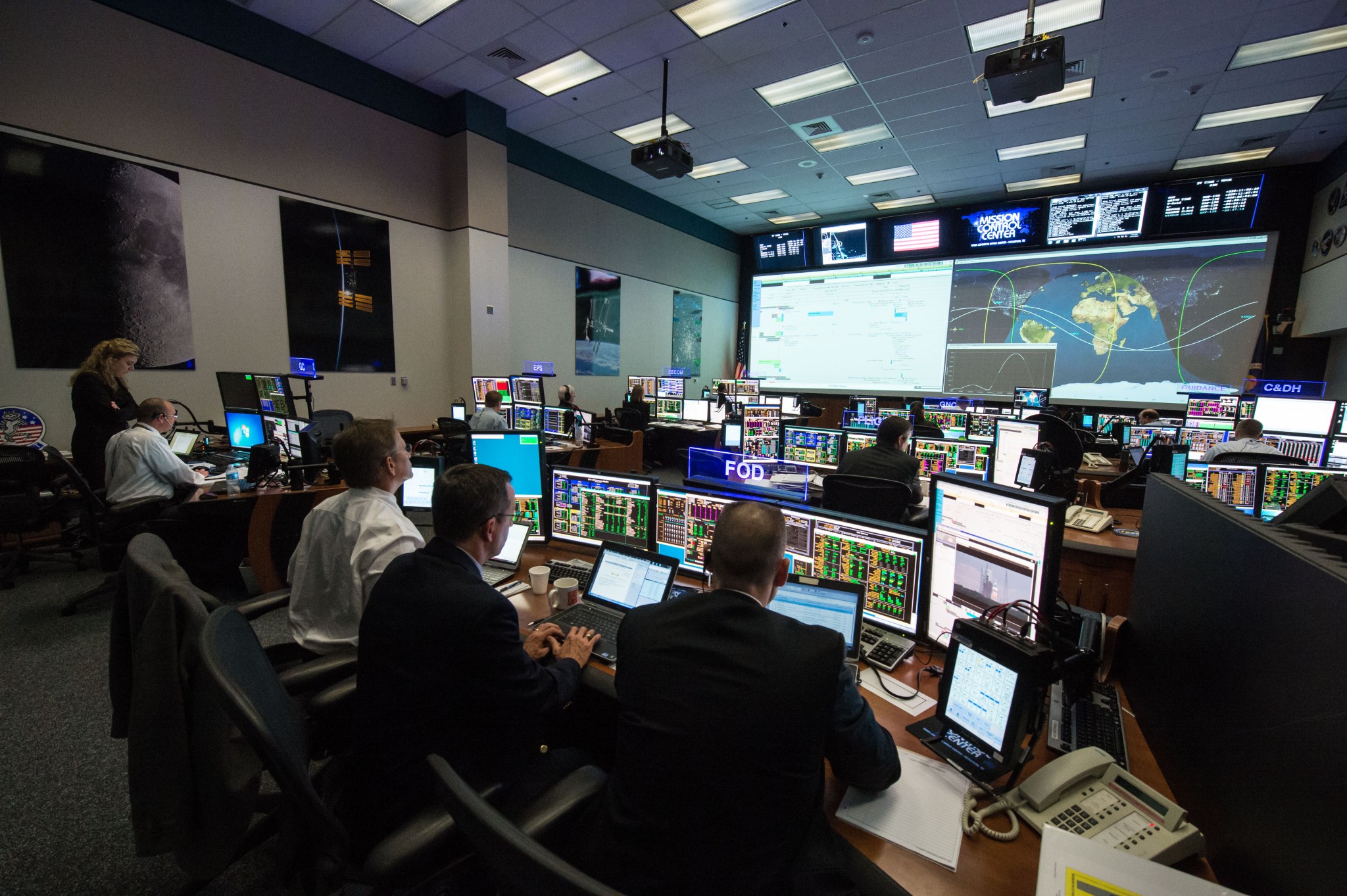 A group of employees sit around individual monitors in an office. The office has a large screen on the far wall with images projected on it. The screen shows a map of the earth. The employees are all focused.