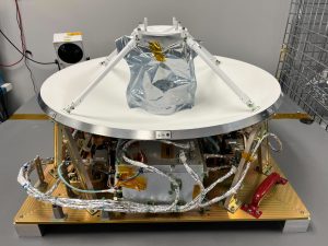 The Polylingual Experimental Terminal (PExT). The terminal’s antenna is protected by a silver thermal sheet, its white parabolic dish supported by four white struts. Aluminum-wrapped cables, metal hardware, and a red carrying handle are mounted on the payload’s gold base.