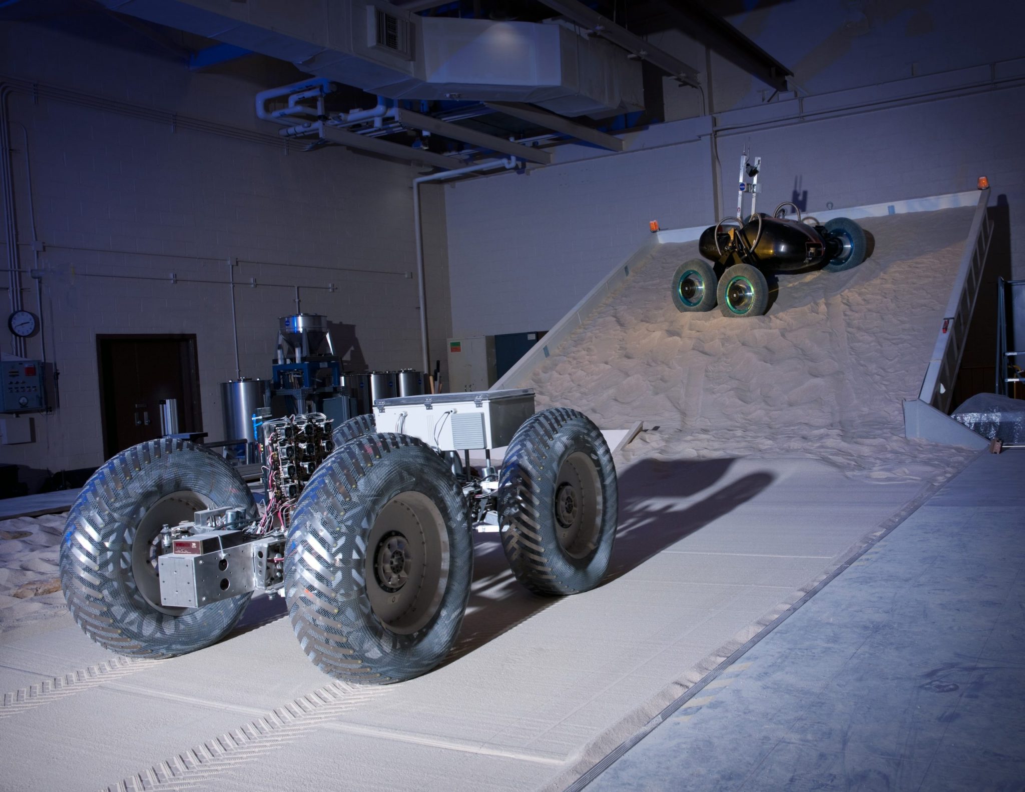 Two test vehicles driving on GRC-1 lunar simulant in the Simulated Lunar Operations Laboratory, one on a flat surface and one on the adjustable tilt-bed