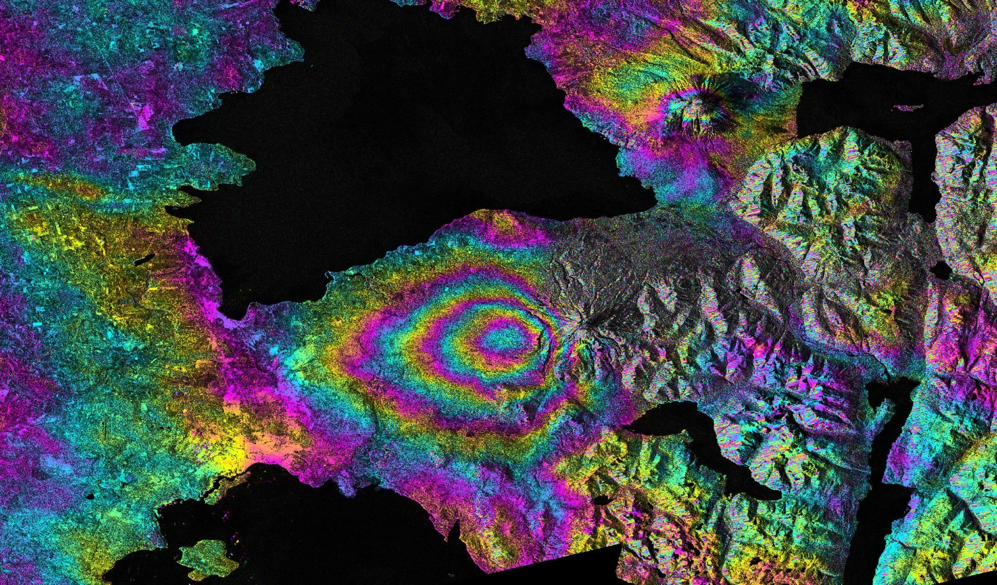 A vibrantly colored interferogram showcasing showing land deformation due to the 2015 eruption of the Calbuco volcano in Chile. The image uses a psychedelic color palette to the color bands west of the volcano to indicate land sinking.