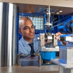 A man wearing glasses operates scientific equipment in a laboratory, adjusting a metal component under a machine surrounded by metal columns and electronic devices.