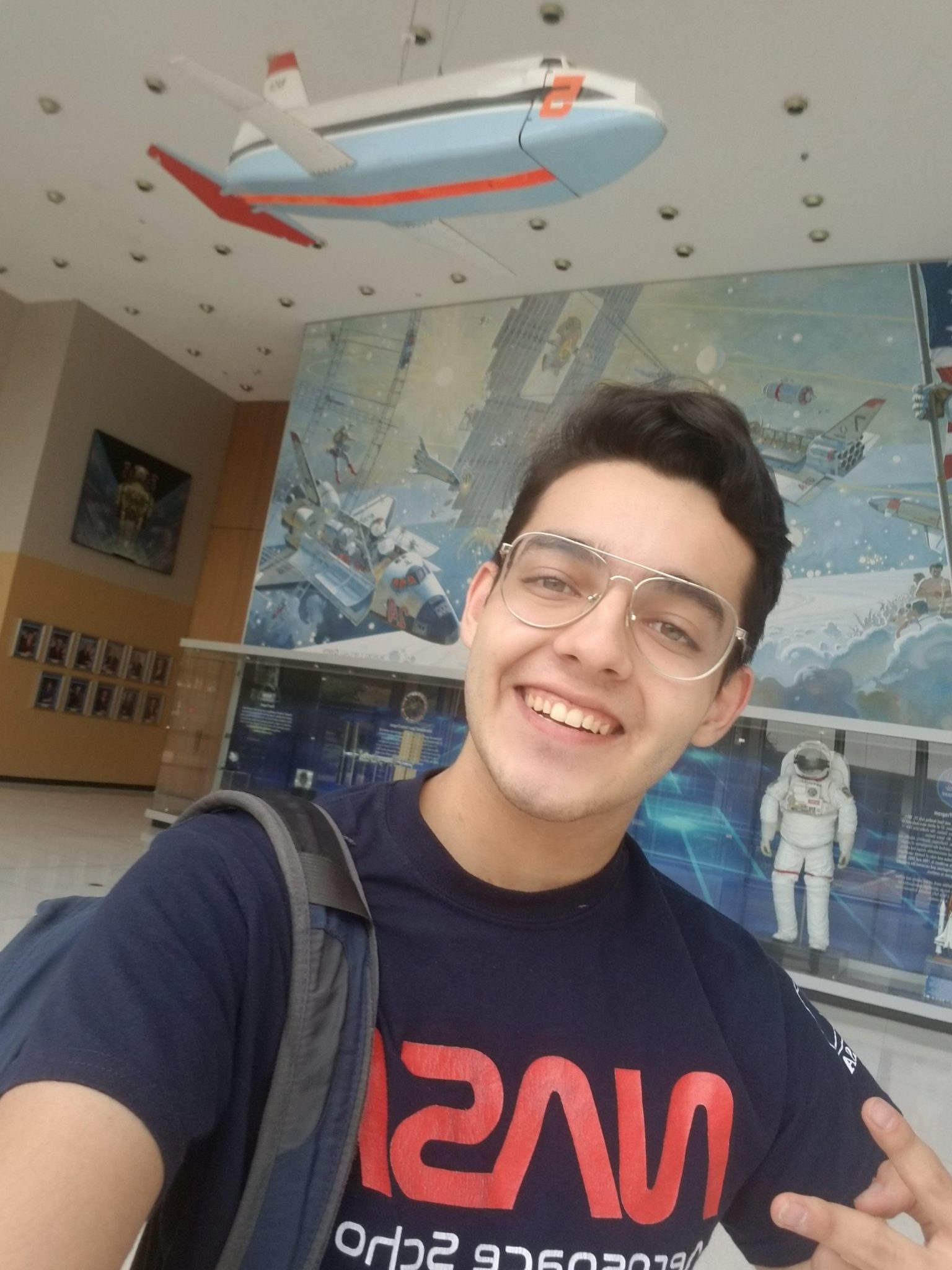 A young man wearing glasses and a NASA t-shirt smiles for a selfie inside a space-themed exhibit, with a suspended model airplane, astronaut suit display, and colorful murals in the background.