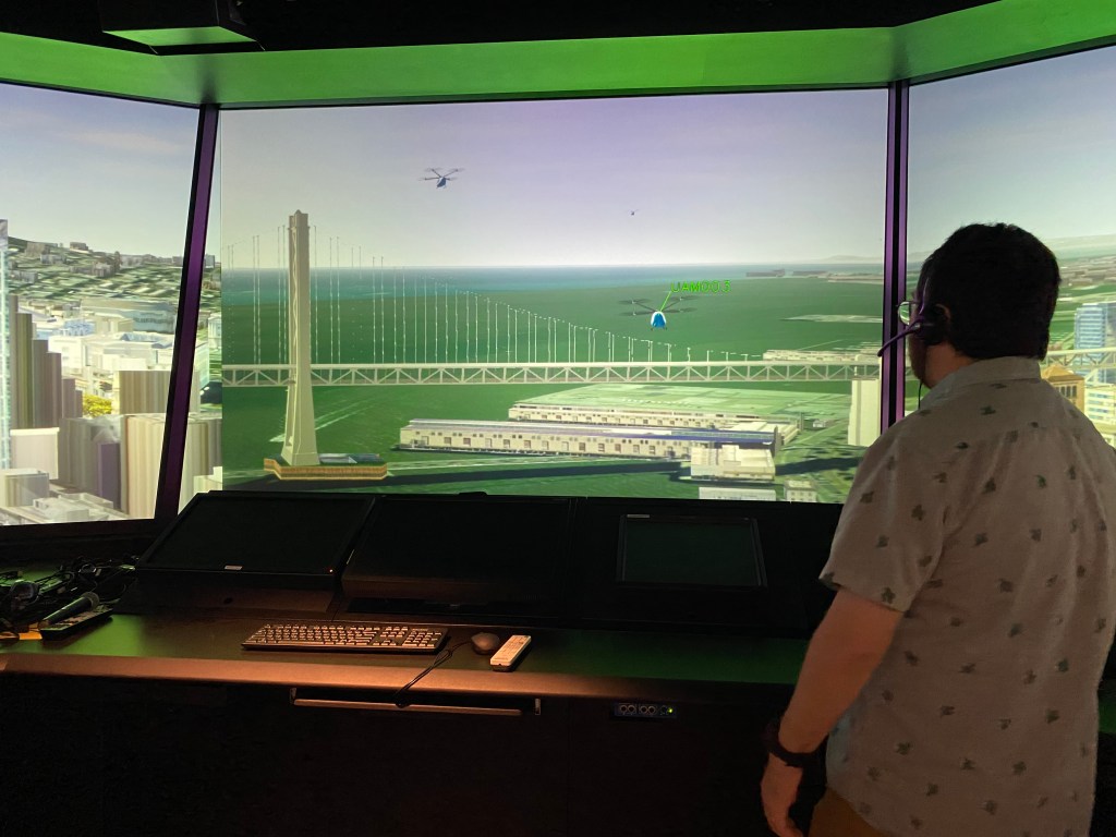 A person wearing a headset monitors the out-the-window views of the FutureFlight Central virtual air traffic control tower, which show two UAM aircraft flying over the San Francisco-Oakland Bay Bridge.