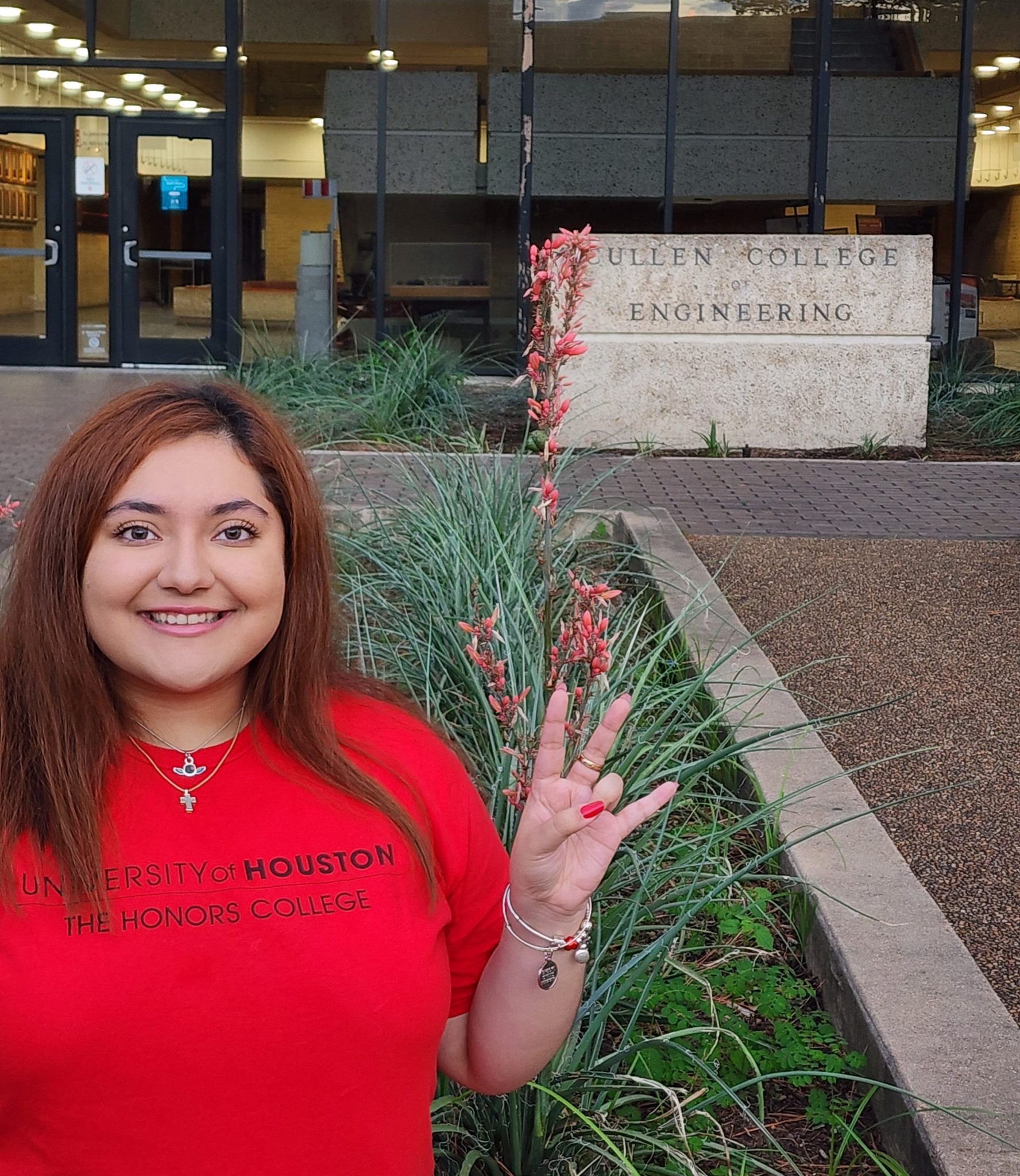 A woman wearing a red ‘University of Houston The Honors College’ t-shirt smiles and holds up a hand gesture in front of the Cullen College of Engineering building, with plants and flowers in the foreground.