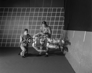 In this black and white photo, two people adjust a 1960s-era spacesuit on a person who is suspended by wires. This third person is parallel to the floor, with his feet on the wall on the right.
