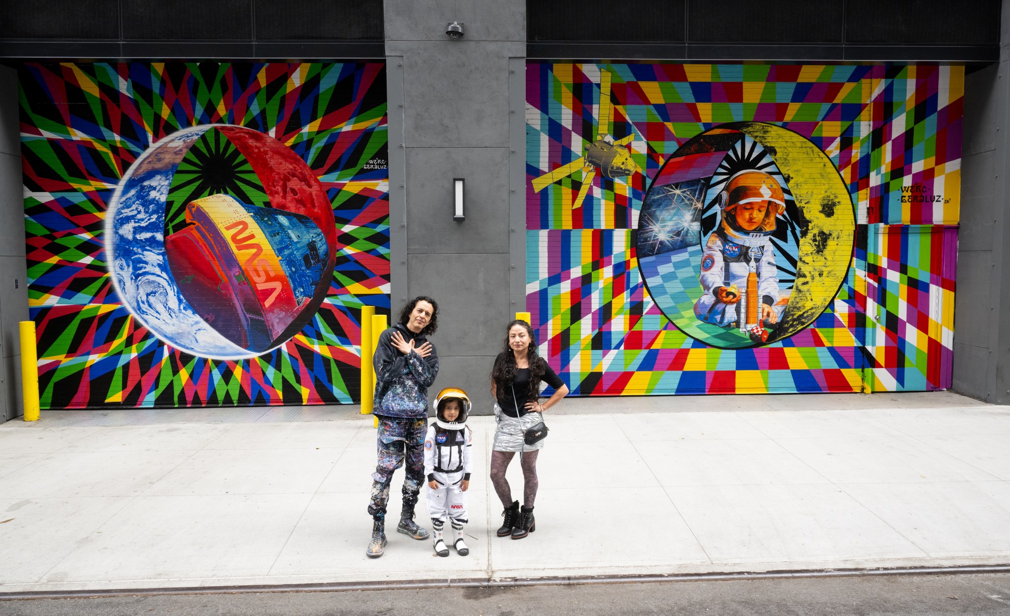 Two adults and a child pose for a photo in front of a colorful space-themed mural. The child wears an astronaut costume.