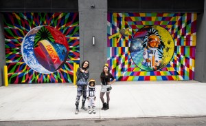 Two adults and a child pose for a photo in front of a colorful space-themed mural. The child wears an astronaut costume.