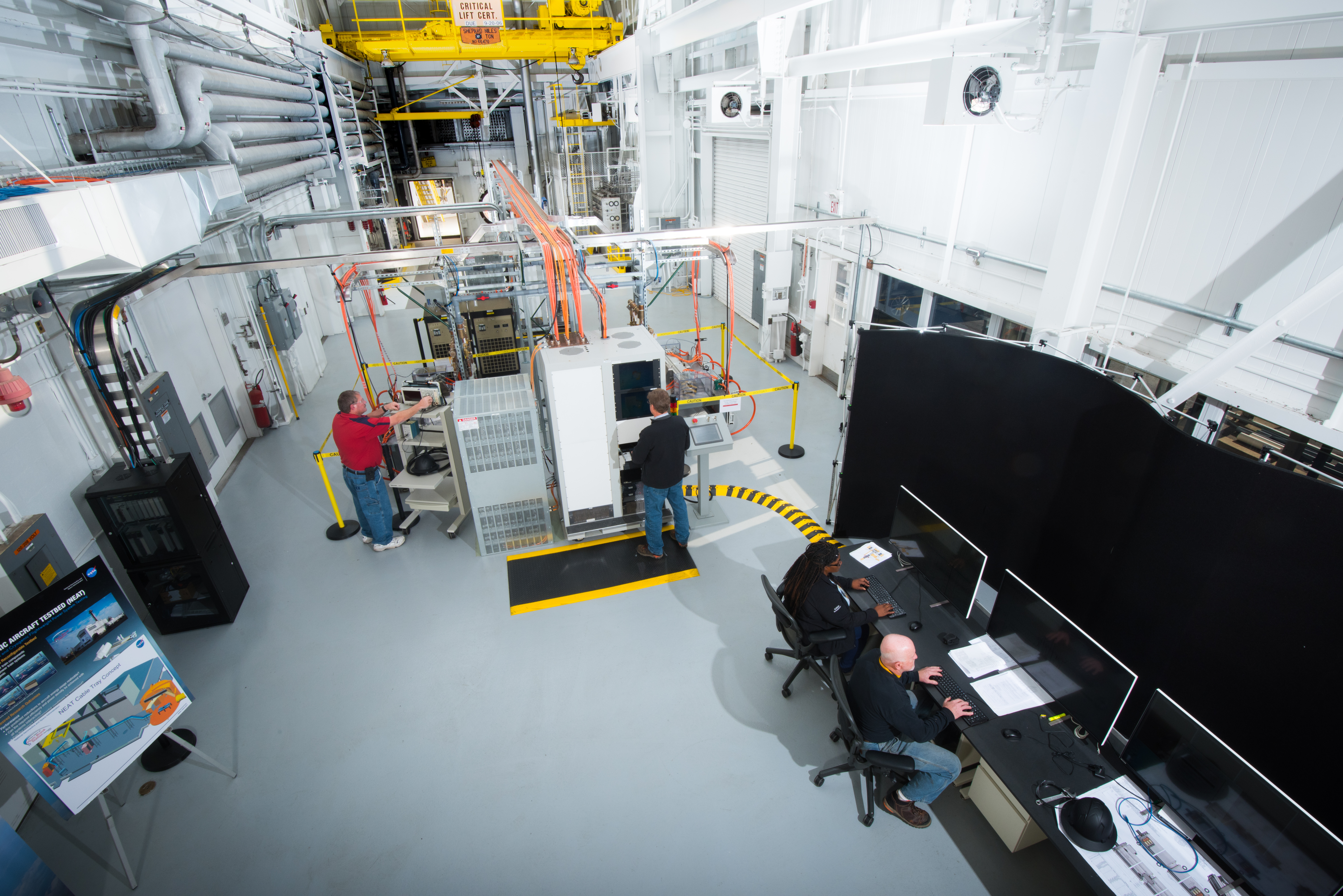A top-down view of a large test building housing various monitors, desks, machinery, and cables. Two people sit at a desk monitoring data on comput...