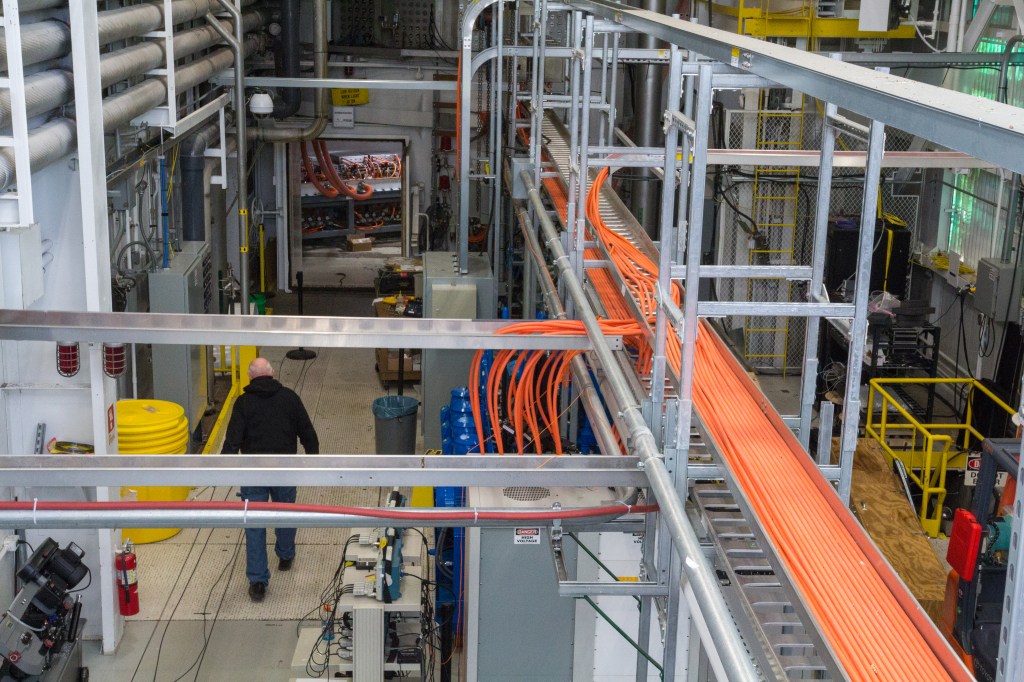 A man walks inside a large test room surrounded by hardware, cables, and metal equipment.