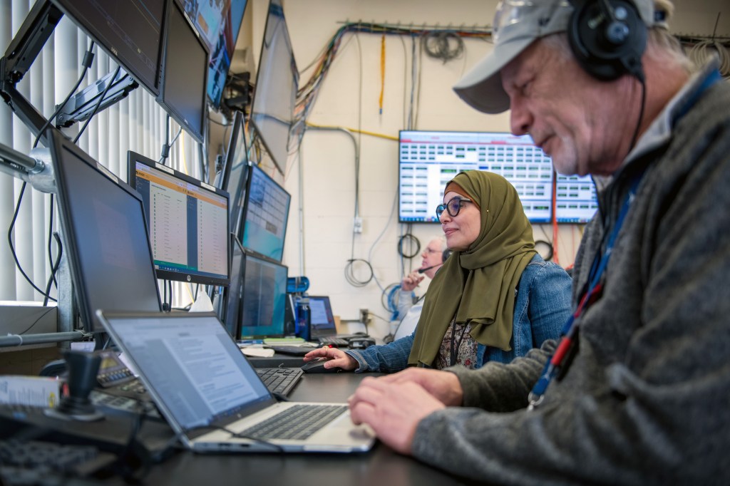 Two men and a women sit at a desk surrounded by laptops and video monitors displaying testing data.