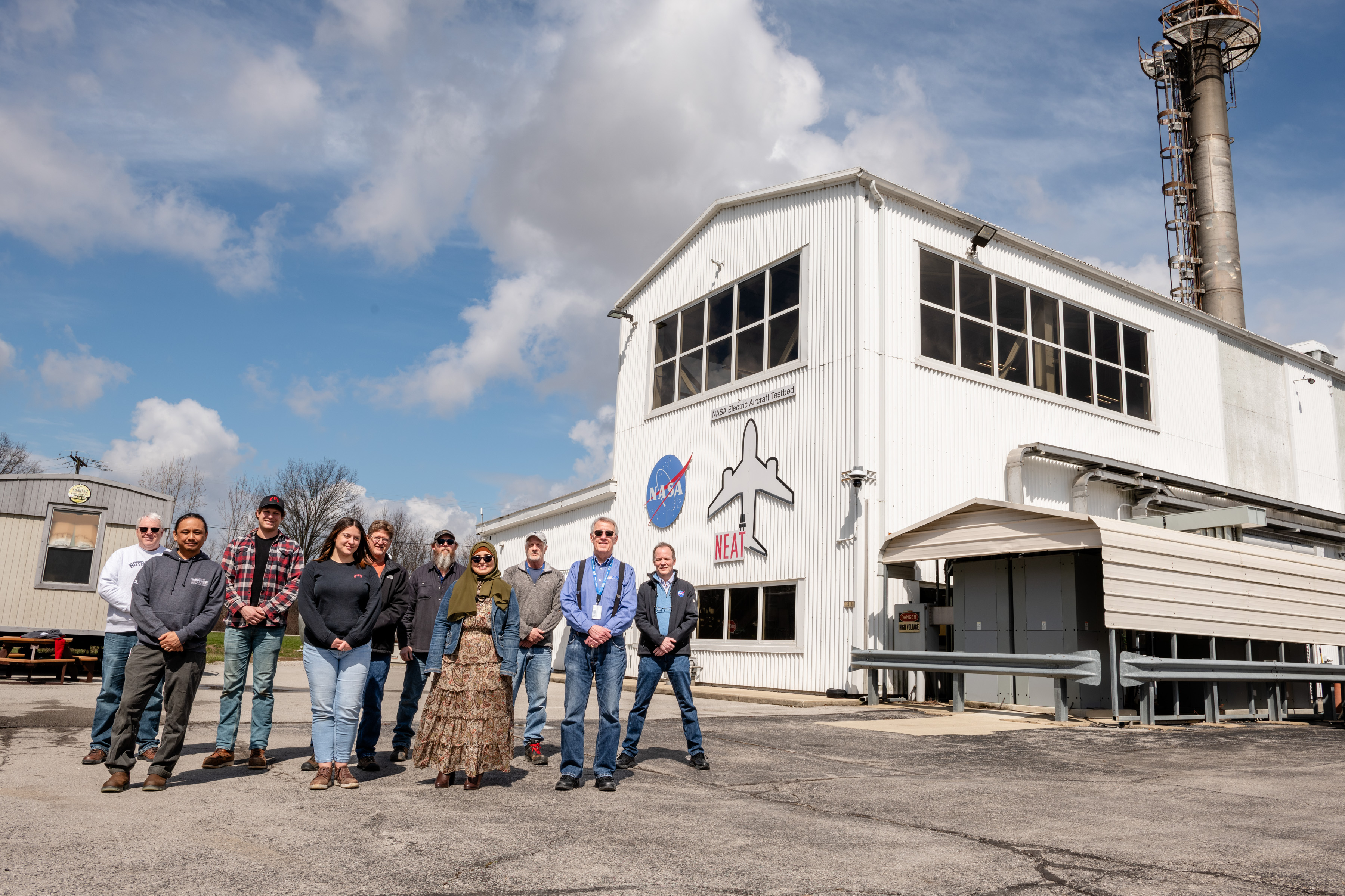 10 researchers from NASA and magniX pose outside of the NEAT facility. The large, white building is seen on the right under a blue sky with clouds ...
