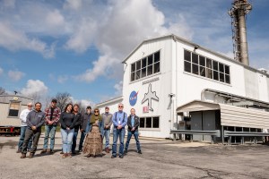 10 researchers from NASA and magniX pose outside of the NEAT facility. The large, white building is seen on the right under a blue sky with clouds in the background.