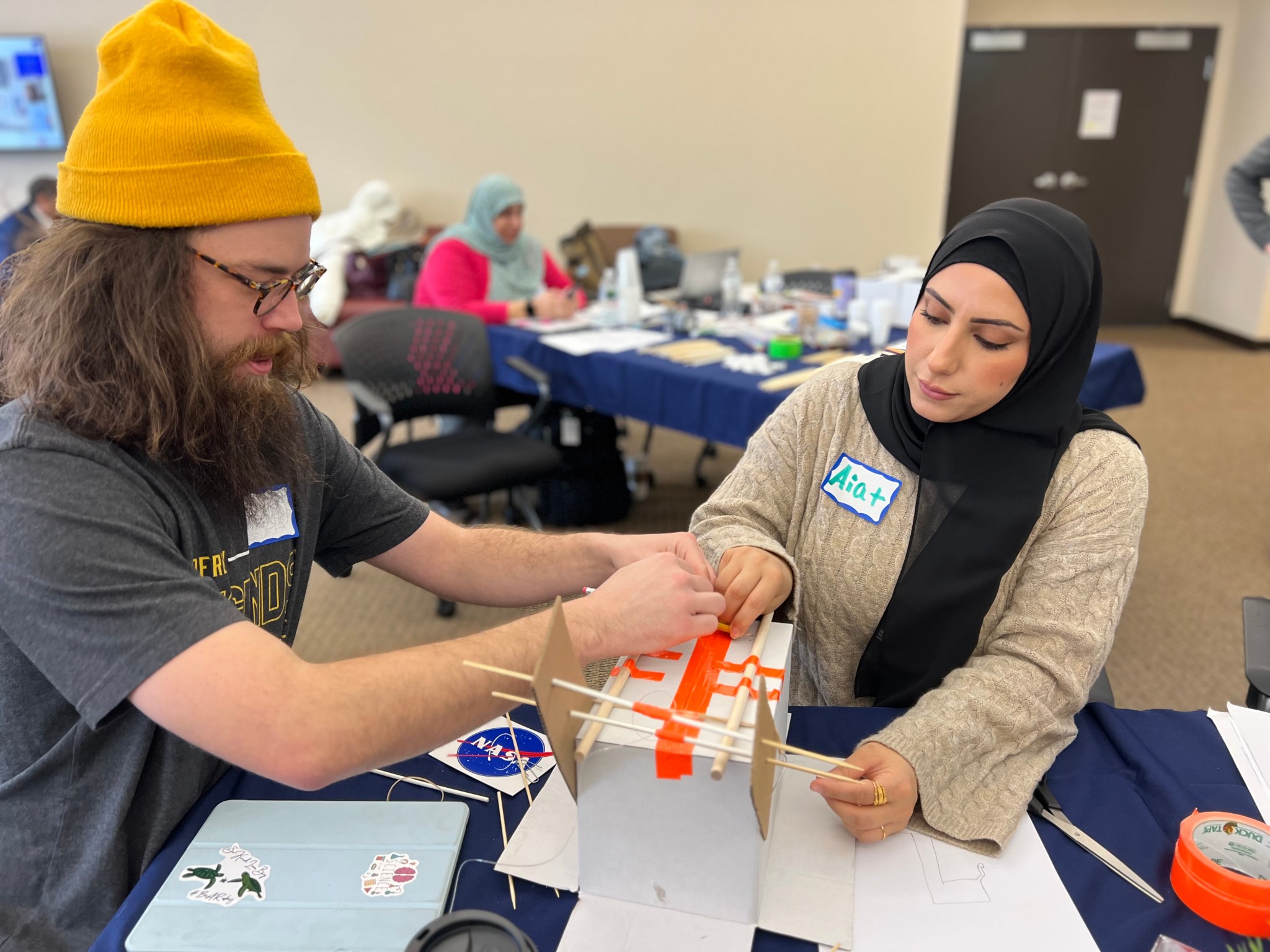 Two after-school educators work with a box, rubber bands, and tape to build a crawler-transporter that will be used for a teaching activity.