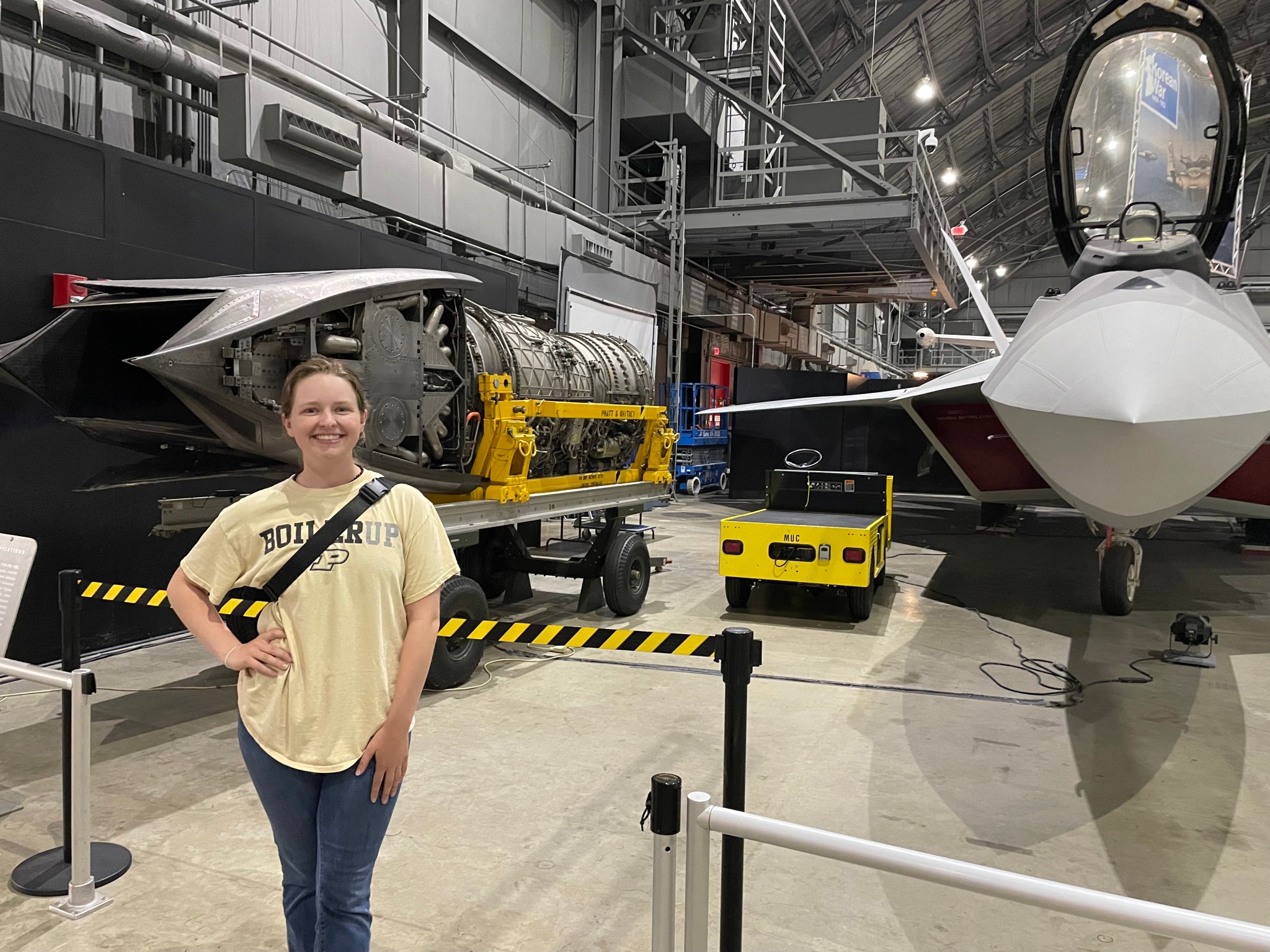 A smiling woman wearing a ‘Boiler Up’ Purdue t-shirt stands inside an aircraft hangar. Behind her are a jet engine on display and a sleek fighter jet.