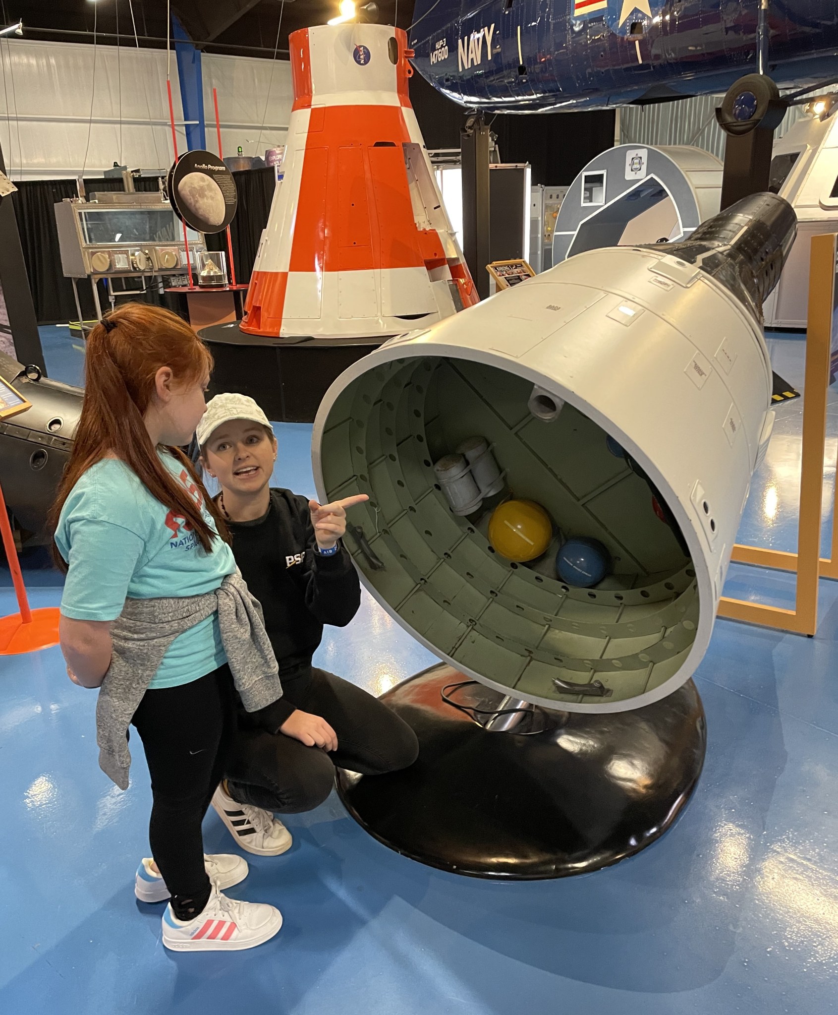 Two individuals, a woman crouching and a young girl standing, engaged in conversation near a space capsule exhibit in a museum.