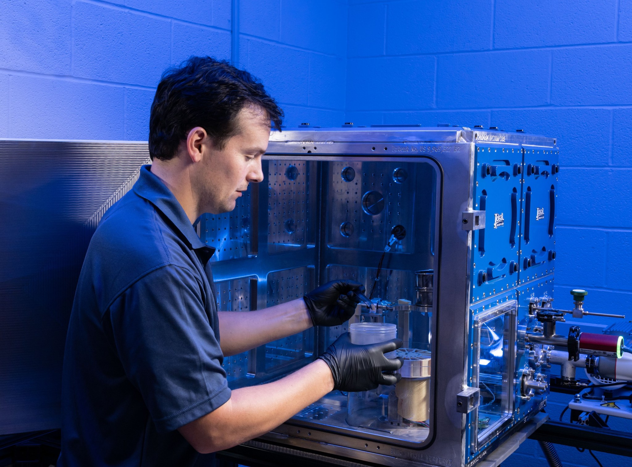 A man with short dark hair, wearing a navy-blue polo shirt and black gloves, works inside a metallic, box-shaped testing chamber with blue panels and multiple knobs, ports, and dials. He is scooping material from a small container.