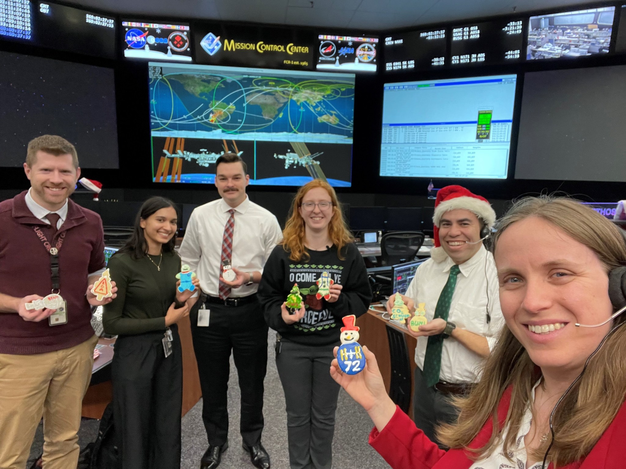 Six flight controllers stand in Johnson Space Center's Mission Control Center, holding up holiday cookies.