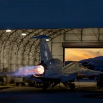 NASA’s X-59 sits partially in a white hangar and emits bright flames with visible shock diamonds from its engine at sunset during maximum afterburner testing outside Lockheed Martin’s Skunk Works Facility in Palmdale, California.