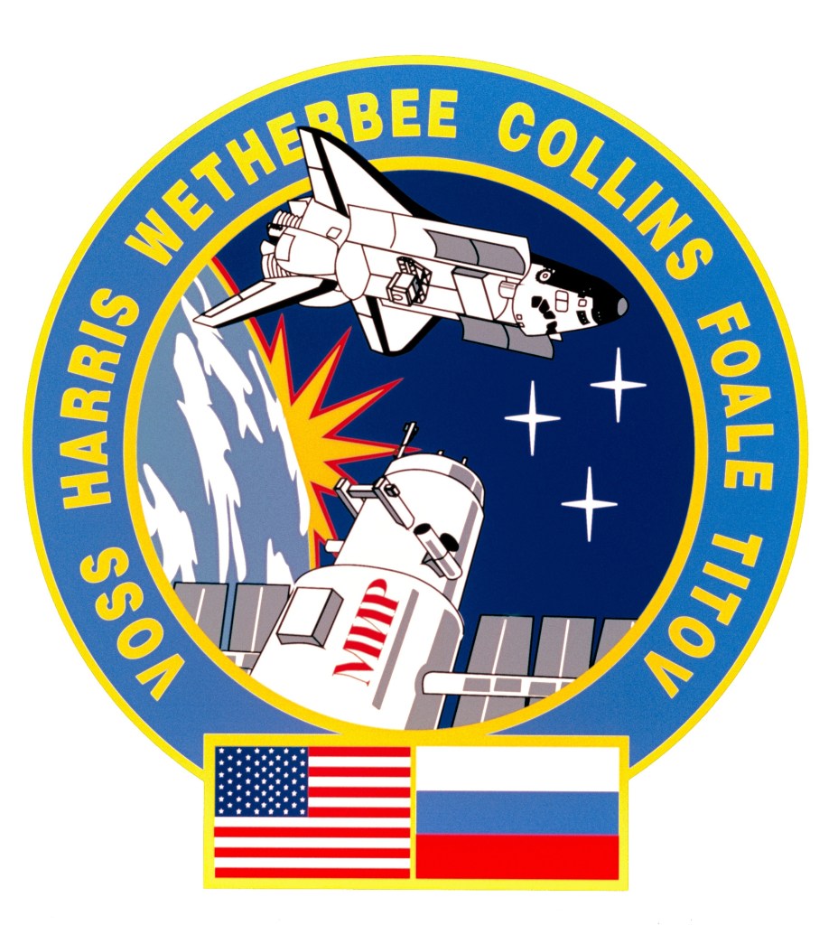 Mostly round mission patch showing a partial Mir space station and a nearby space shuttle, a US and Russian flag at the bottom and the names of the 6 astronauts around the edge of the patch.