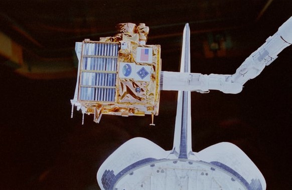Image of a box-shaped gold and blue satellite, grappled by a robotic arm, suspended in front of the tail of the space shuttle.