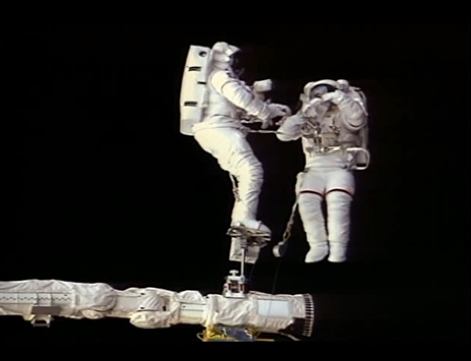 Image of two spacewalking astronauts in white spacesuits against the blackness of space.