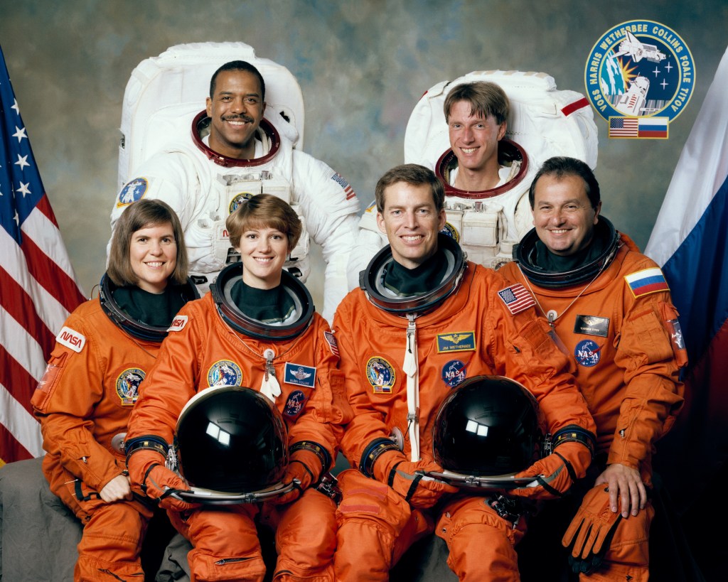 A group photo of six astronauts, four in the front row wearing orange space suits and two in the back row in white spacewalking spacesuits