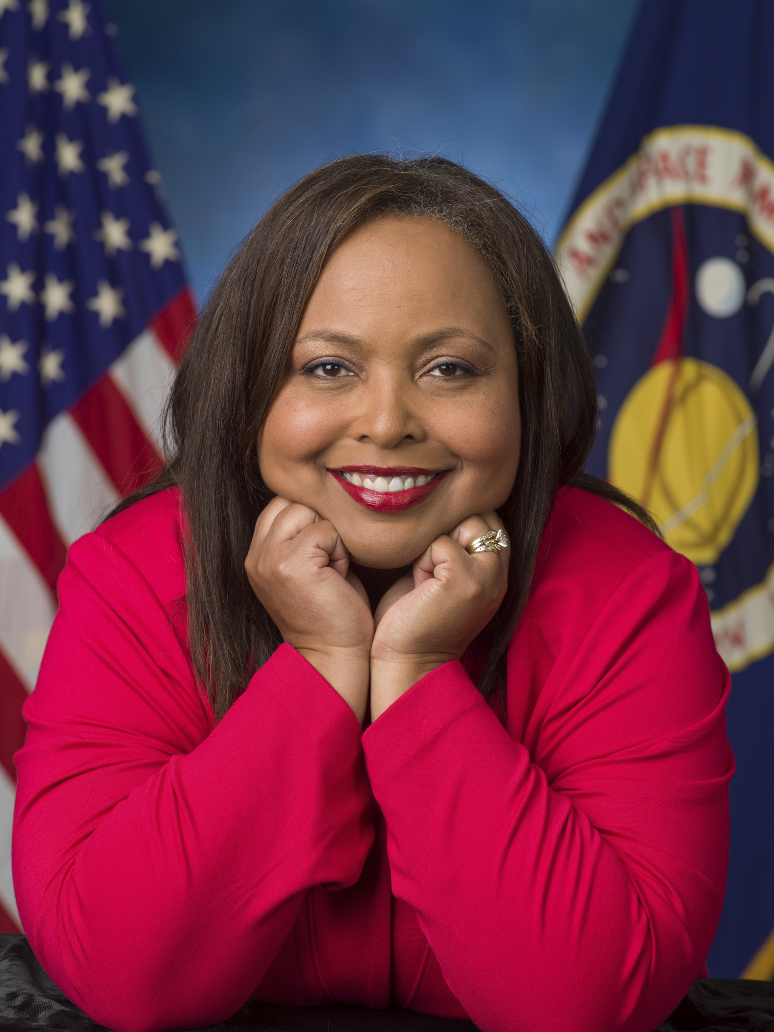 A woman wearing a bright red blazer is smiling with her chin resting on her hands. She is posed in front of a backdrop featuring the U.S. flag and a NASA logo.