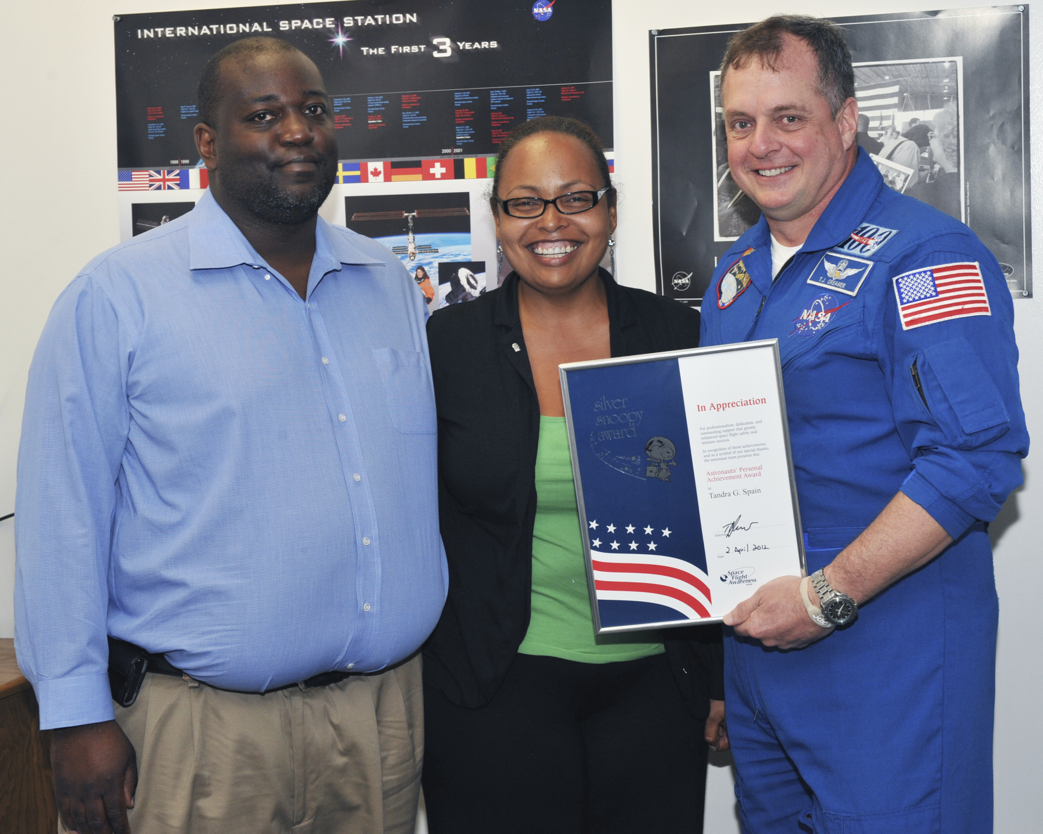 Three people are standing together, smiling. Behind them is a poster featuring the International Space Station and several international flags.