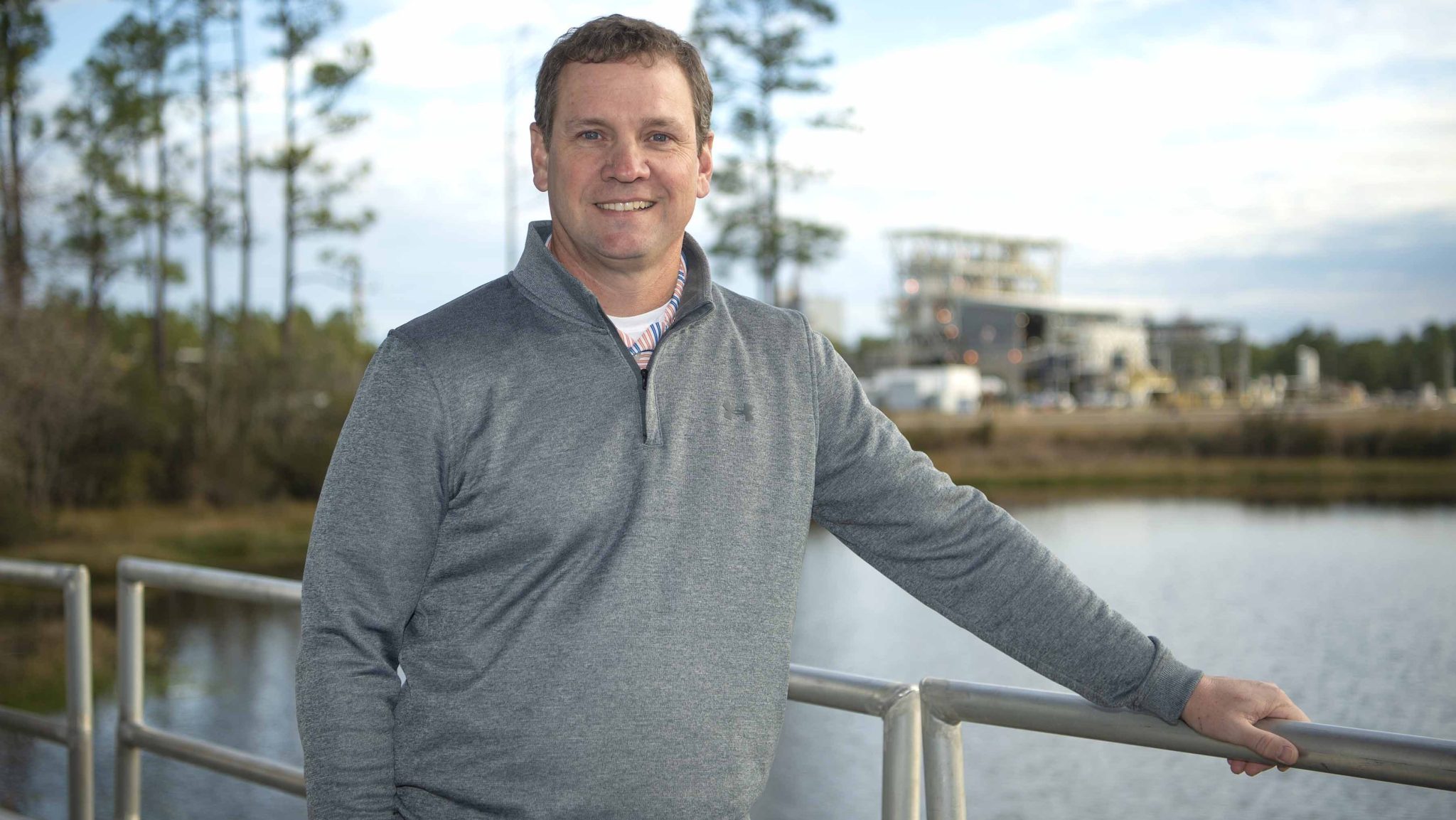 Tim Stiglet stands for a portrait wearing a gray pullover; A Stennis utility facility is shown in the background