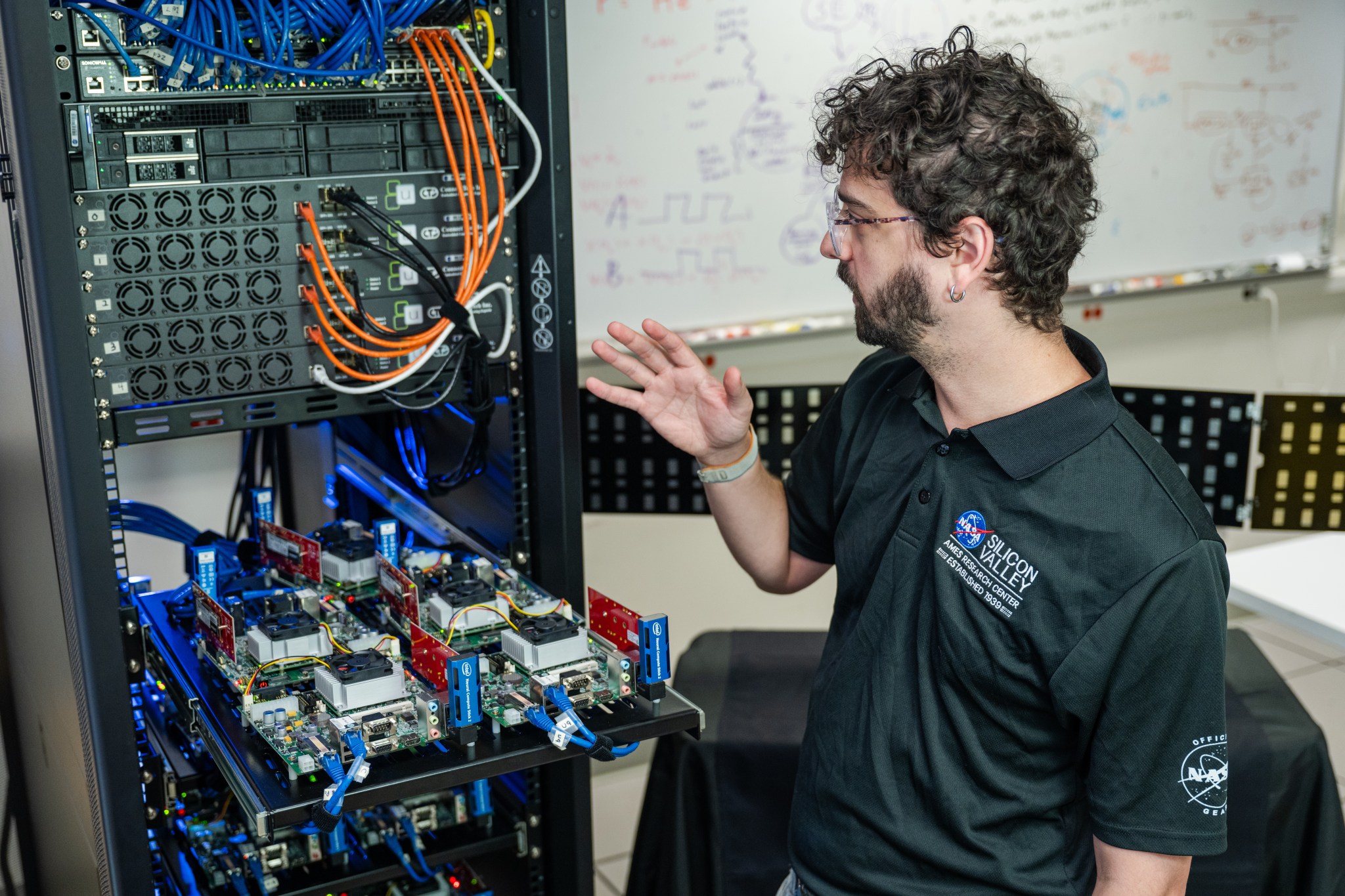 A man stands in front of a computer server and gestures towards the racks and cables.