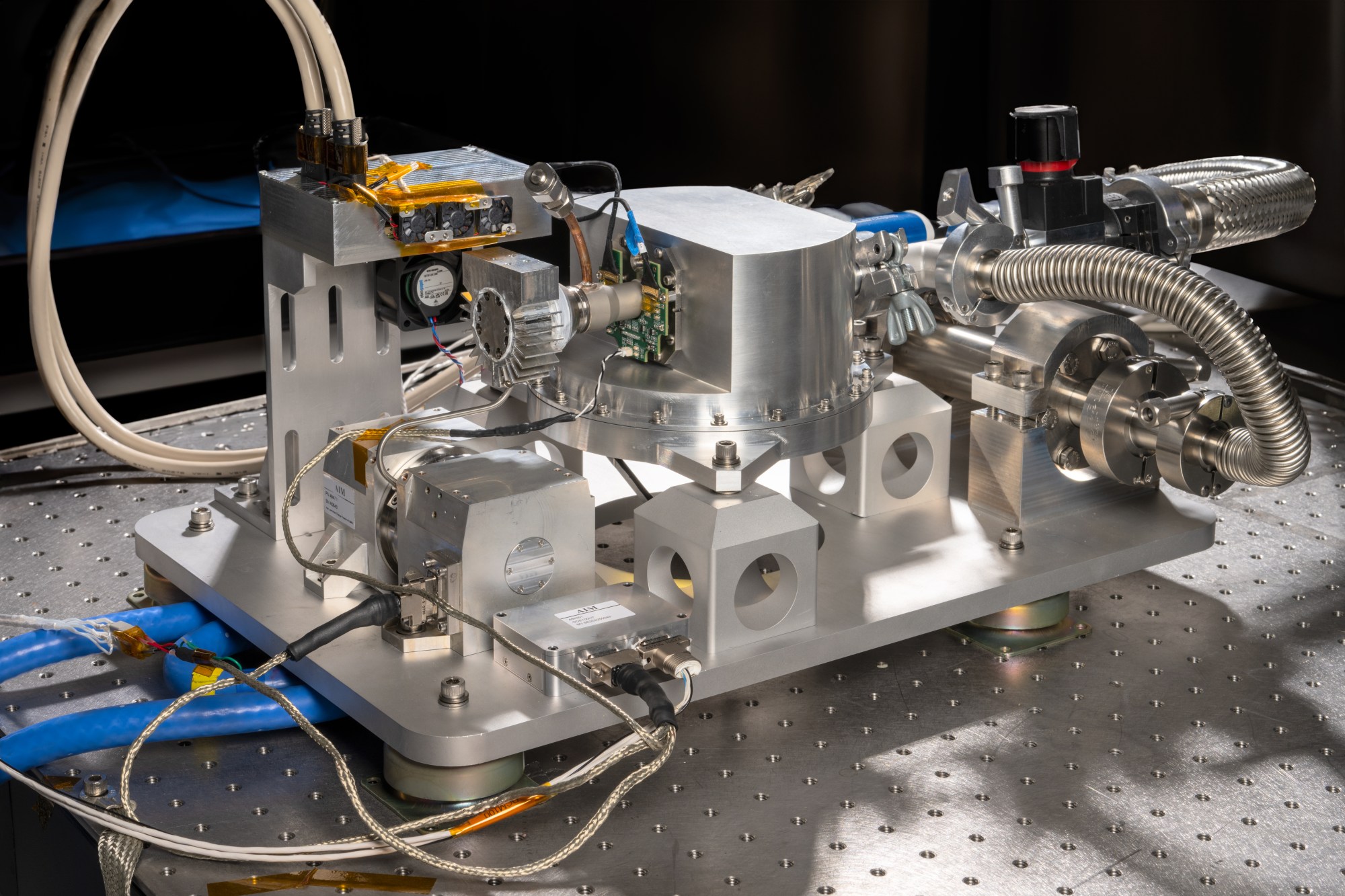 A scientific instrument sits on top of a metal surface. The instrument is composed of several metal blocks, cables, tubes, and dials. The central block appears to have a microchip and is connected by cables and tubes to the adjacent blocks. Some electrical wiring protrudes from parts of the instrument.