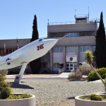 A large white airplane on a stand sits in desert plant and stone landscaping in front of an of a two-story office building.