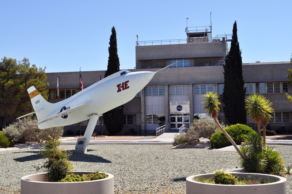 A large white airplane on a stand sits in desert plant and stone landscaping in front of an of a two-story office building.