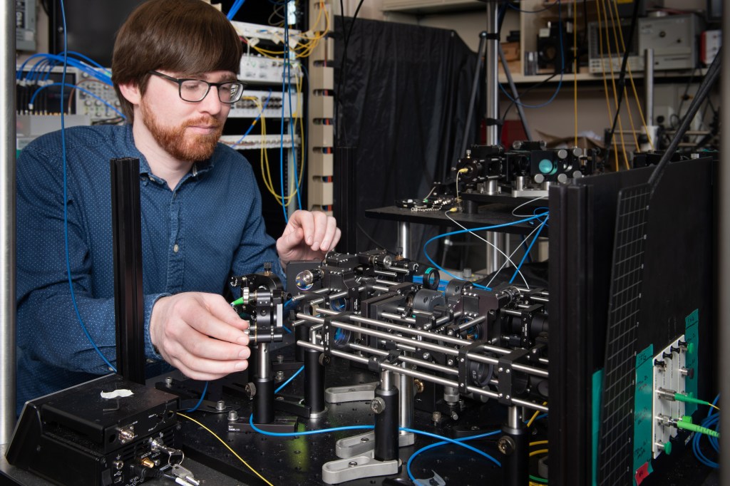 A person with glasses and a beard works on a complex setup of electronic and optical equipment in a laboratory. There are various wires and components on the table. They are wearing a blue shirt and focusing on adjusting the devices.