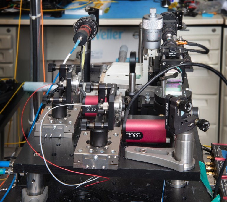 Close-up of a complex electronic device setup, featuring various metal components, wires, and lenses arranged on a black platform. Instruments and cables are connected, suggesting scientific or industrial use. Background shows a cluttered lab bench.