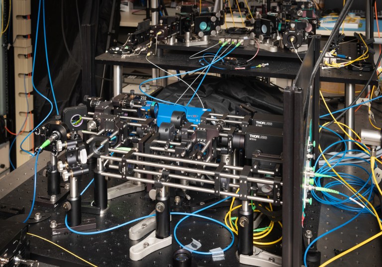 A complex laboratory setup featuring multiple optical devices and electronic components, interconnected with blue and yellow cables, likely part of a scientific experiment or research project. Shelves with additional equipment are visible in the background.