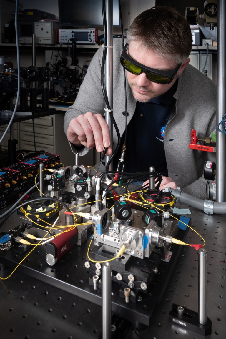A person wearing protective sunglasses operates complex optical equipment in a laboratory. The setup includes various lenses, mirrors, and electronic components on a perforated table. The person is adjusting a component with precision.