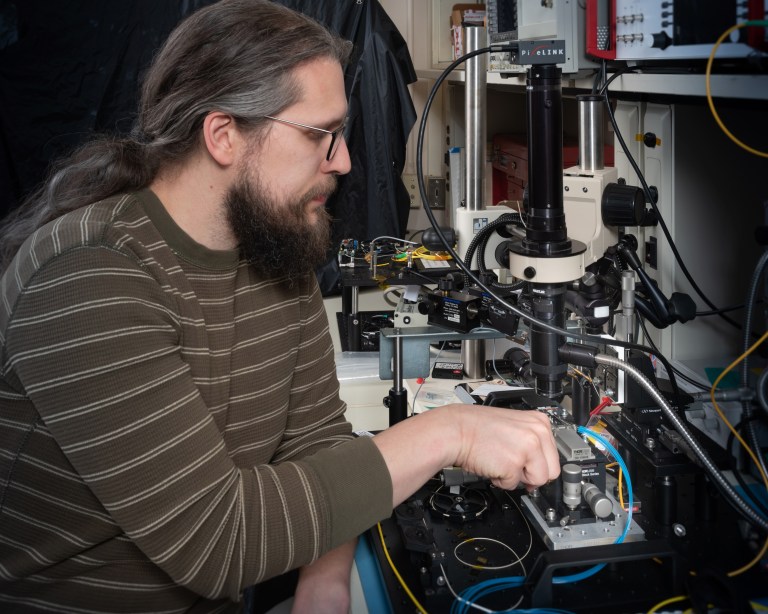 A person with long hair and glasses operates a complex electronic testing device in a lab. The equipment has multiple wires and components. The individual is focused on adjusting a part of the machinery.