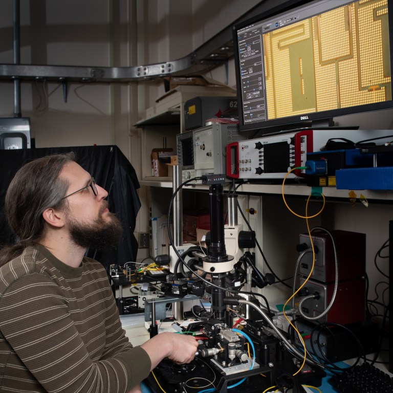 A man with long hair and glasses operates electronic equipment in a lab. He is focused on a computer screen displaying a grid pattern. Various wires and devices surround him in the workspace.