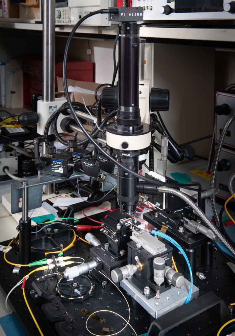 A complex scientific setup featuring a microscope with several attached wires, cables, and components. The equipment is placed on a cluttered lab bench surrounded by various scientific instruments and lab tools.