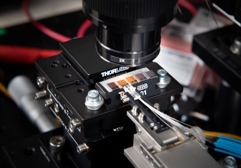 Close-up of scientific equipment showing a microscope lens positioned above a mechanical stage. Several metallic and electronic components are visible, including wires and circuit boards. The setup appears to be part of a laboratory experiment.