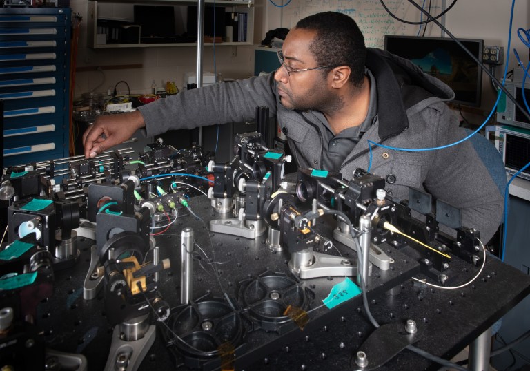 A person in a lab coat is adjusting equipment on a cluttered optical table filled with lasers and scientific instruments. The room has shelves and a whiteboard in the background. Cables are visible hanging above.